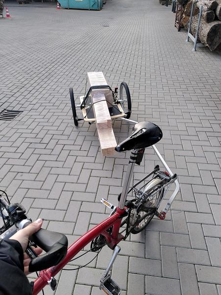 A bike trailer loaded up with a stack of cling wrapped wood. The trailer
is hitched to the back of a black and red Brompton, most of which is
visbible in the foreground
