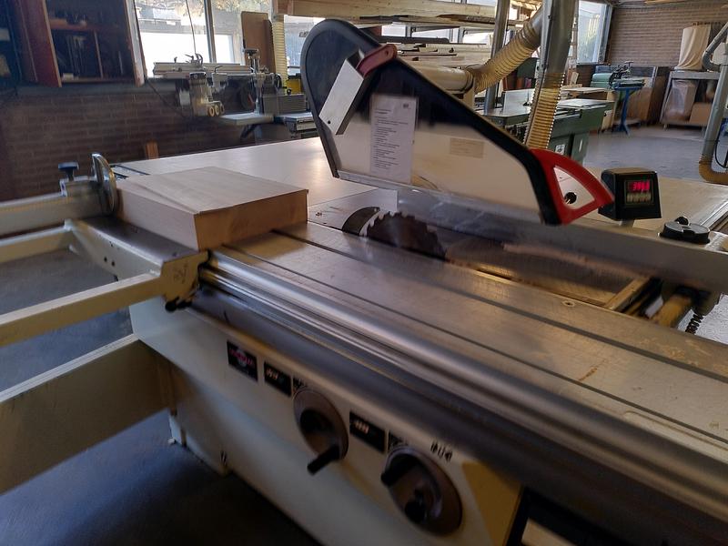 A large sliding table cabinet saw with a block of beech sat on it just
past the blade. A sliver of beech is sat on top of the block, it's just
been cut off the main block