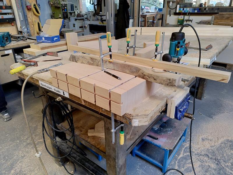 A block of beech clamped to a workbench by a clamp on the underside with
a yellow and green handle. The block has a horizontal groove across the
front, with five more vertical grooves on the same face. The top has two
grooves running across it. There's two pieces of wood clamped to the top
of the block. Behind the block a bluey green Makita Router is visible. The
floor is covered in saw dust.