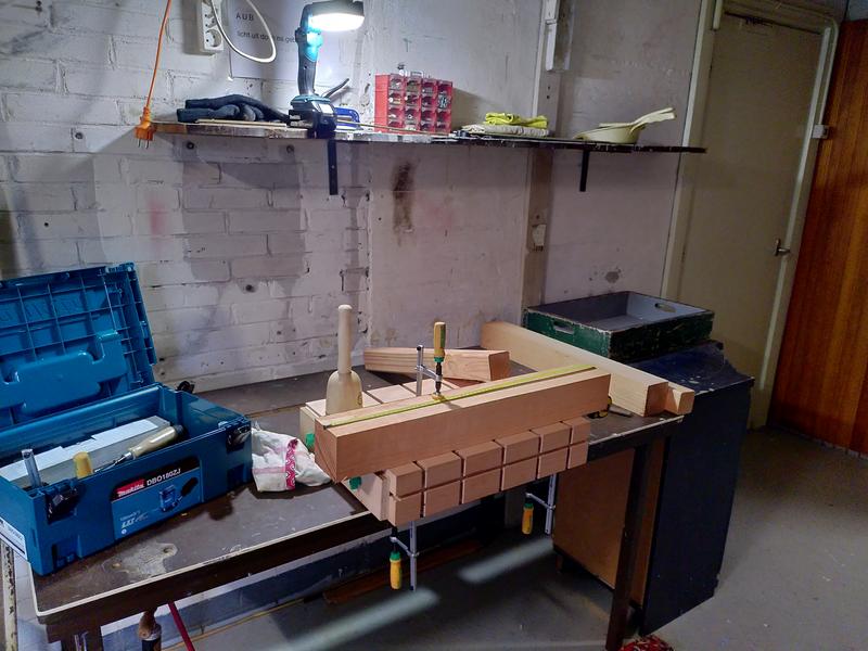 A workbench made of a block of beech, clamped to a table. The block has
a number of channels routed into it. On top of the block is a length of
square section douglas fir. The douglas fir is clamped to the beech block
using a clamp with a yellow and green handle. A wooden mallet is next to
the douglas fir, a tape measure is running across the top. A bluey green
Makita tool box is open to the left of the beech work bench. A few more
lengths of douglas fir surround the bench.