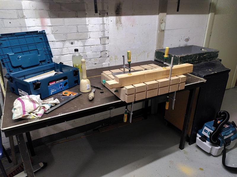 A workbench made of a block of beech clamped to a table. On top of the
bench is a length of douglas fir. A half lap joint has been cut into one
end of the douglas fir. It's clamped in place with two clamps with yellow
and green handles. On the floor to the right sits a makita vacuum cleaner.
On the table to the left is an open makita tool box, a bottle of lemonade,
a wooden mallet, a chisel, and a combination square.