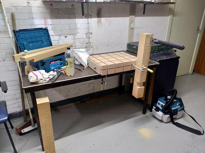 A table covered in wood working tools and wood. On the right is a work
bench made of a block of beech, it's clamped to the table. A large square
section of douglas fir is clamped to the front of this workbench. It has a
half lap joint cut near one end, and a japanese saw is sticking out of a
cut in the top of it. On the left of the table a makita tool box is open,
with some more douglas fir sitting on top of it, along with a bottle of
lemonade, a combination square, and a white plastic bag. On the floor
another length of douglas fir, and a makita vacuum cleaner.