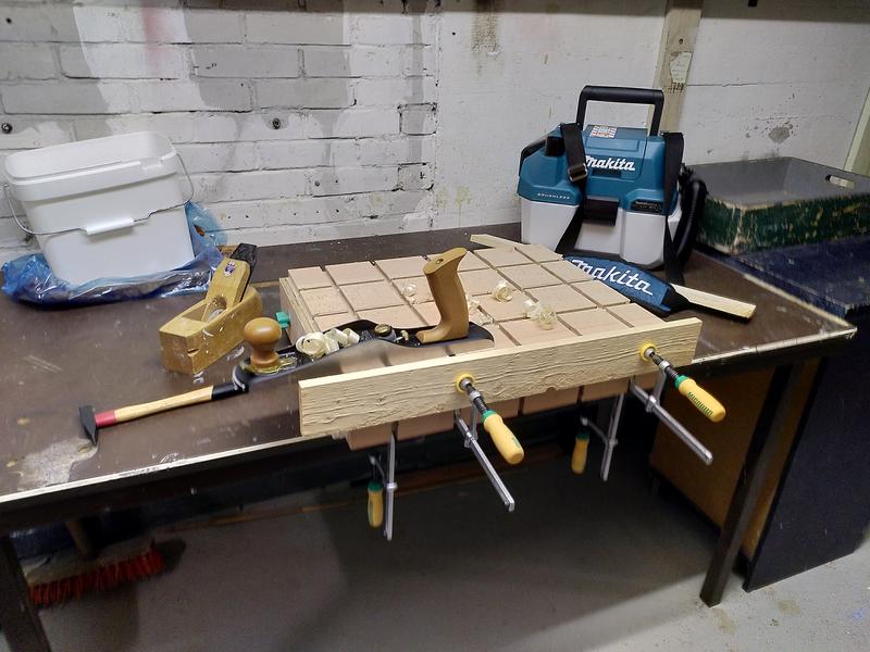 A workbench made of a block of beech clamped to a table. A peice of pine
wood is clamped to the front of the bench using a pair of clamps with
yellow and green handles. On top of the bench sits a metal low angle jack
plane and a few wood shavings. To the right is a makita vacuum cleaner. To
the left a small hammer, a wooden coffin smoother hand plane, and a white
bucket.