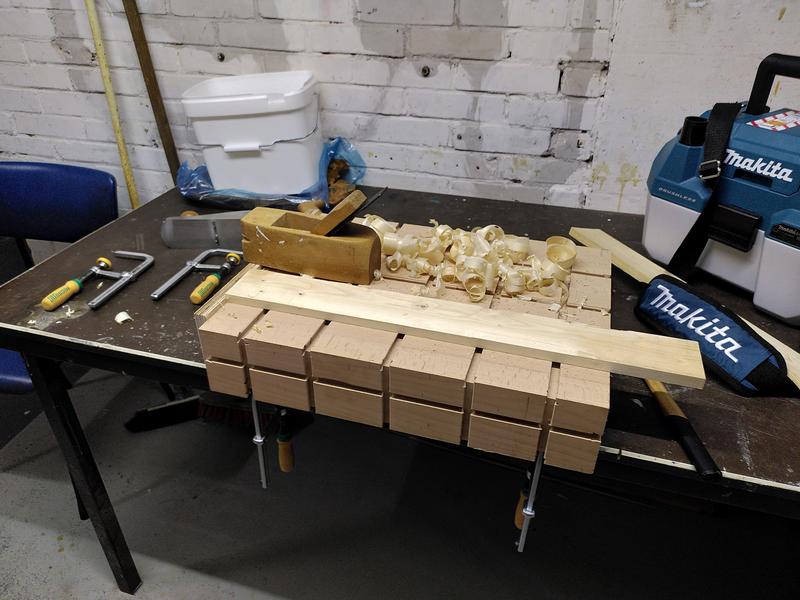 A Workbench made of a block of beech clamped to a table. The block has a
number of channels routed into it. On top of the block is a think piece of
pine wood. This is butted up against a planning stop on the end of the
beech block. A pile of wood shavings and a wooden coffin smoothing plane
sit next to the pine wood. A makita vacuum cleaner is to the right of the
workbench. To the left are two clamps with yellow and green handles.
Beyond those a metal hand plane is laying on it's side in front of a white
plastic bucket.